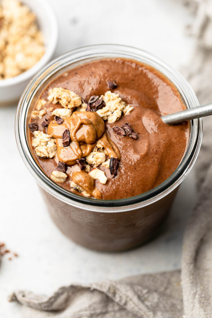 Chocolate post workout smoothie in a large glass jar topped with nut butter, granola, and cacao nibs. A spoon is coming out of the jar. Behind the jar is a bowl of more granola.