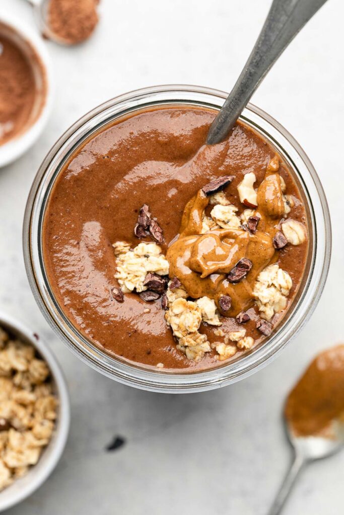 Overhead shot of a chocolate protein shake topped with granola and nut butter with a spoon stuck in it. Around the jar is a bowl of granola, cacao powder, and spoon with nut butter on it.