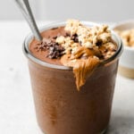 Chocolate protein smoothie in a glass jar with a spoon in it. It is topped with granola, nut butter, and cacao nibs. Around it is a teaspoon with cinnamon, some granola, and bowl of granola.
