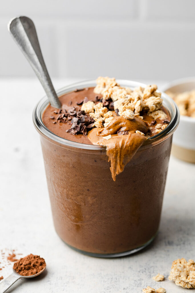 Chocolate protein smoothie in a glass jar with a spoon in it. It is topped with granola, nut butter, and cacao nibs. Around it is a teaspoon with cinnamon, some granola, and bowl of granola.