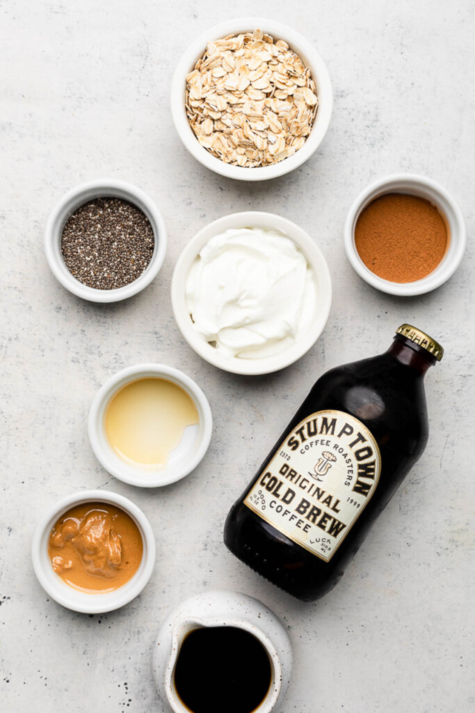 Grey countertop with a bowl of rolled oats, a bowl of cinnamon, a bowl of greek yogurt, a bottle of cold brew coffee, a pitcher of cold brew, a bowl of cashew butter, a bowl of honey, and a bowl of chia seeds.