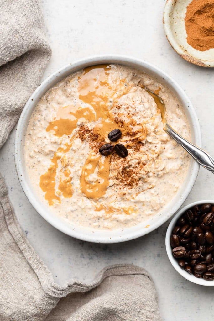 Bowl of coffee overnight oats topped with cashew butter and coffee beans with a spoon in it. Around the bowl is a tan linen, a bowl of coffee beans, and a small bowl of cinnamon.