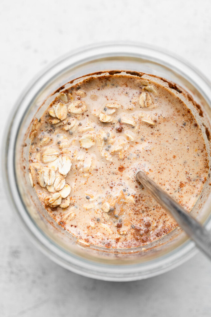 Overhead shot of a jar filled with a liquid mixture of cold brew, rolled oats, cinnamon and chia seeds with a spoon in it.
