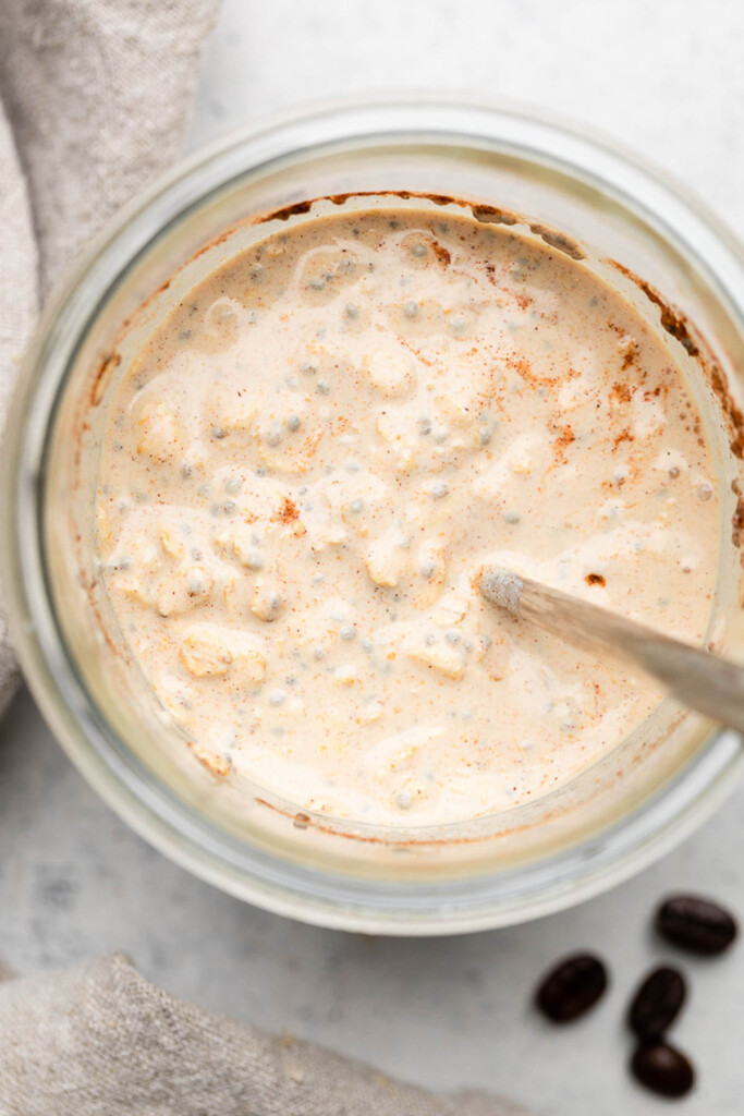 Overhead shot of a jar filled with a creamy mixture of rolled oats, chia seeds, and cinnamon with a spoon in it. Next to the jar is a tan linen and some coffee beans.