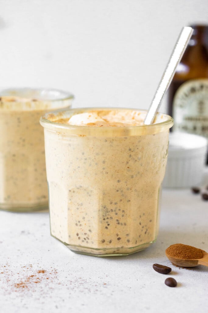 Two jars of overnight oats with coffee, with one being behind the other. A spoon is in the jar in the front. In front of the jar is a teaspoon of cinnamon and some coffee beans. Off to the back is a bottle of cold brew and a small white dish.