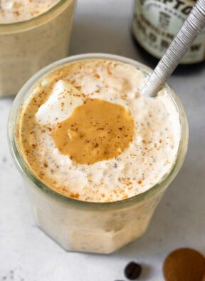 A jar of coffee overnight oats topped with yogurt, cashew butter, and cinnamon with a spoon in the jar. Behind it is another jar of overnight oats and bottle of cold brew coffee. In front of the jar is a teaspoon of cinnamon and some coffee beans.