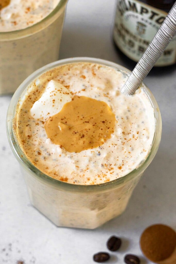 A jar of coffee overnight oats topped with yogurt, cashew butter, and cinnamon with a spoon in the jar. Behind it is another jar of overnight oats and bottle of cold brew coffee. In front of the jar is a teaspoon of cinnamon and some coffee beans.