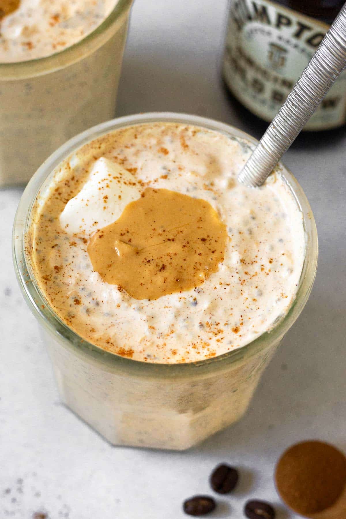 A jar of coffee overnight oats topped with yogurt, cashew butter, and cinnamon with a spoon in the jar. Behind it is another jar of overnight oats and bottle of cold brew coffee. In front of the jar is a teaspoon of cinnamon and some coffee beans.