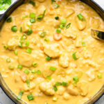 Large pot of vegan buffalo cauliflower potato soup garnished with green onions. Around it is a small bowl of green onions and a bowl of buffalo sauce.