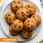 Pumpkin protein balls stacked on top of each other on a small white plate. Off to the side of the plate is a bowl of mini chocolate chips, a bowl of nut butter, and measuring spoons.