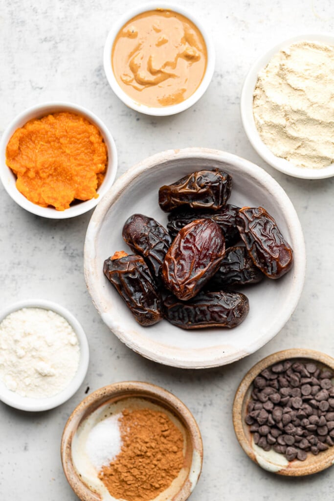 Grey counter with a bowl of nut butter, a bowl of vanilla protein powder, a bowl of medjool dates, a bowl of mini chocolate chips, a bowl of spices, a bowl of flour, and a bowl of pumpkin puree.