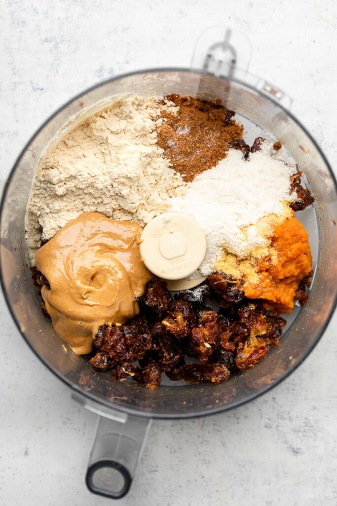 Food processor with broken up medjool dates, cashew butter, vanilla protein powder, spices, coconut flour, and pumpkin puree before it is blended together.