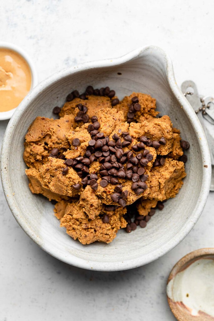 Bowl filled with a ball of dough that has been broken up with mini chocolate chips sprinkled on top. Next to is a bowl of nut butter and a bowl of flour.
