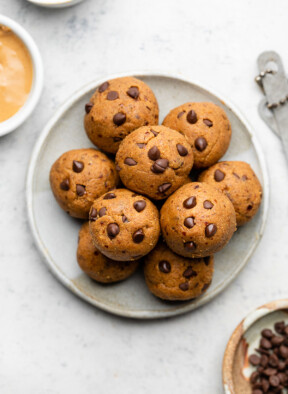 Pumpkin protein balls stacked on top of each other on a small white plate. Next to the plate is a bowl of mini chocolate chips, a bowl of nut butter, a bowl of pumpkin puree, and measuring spoons.