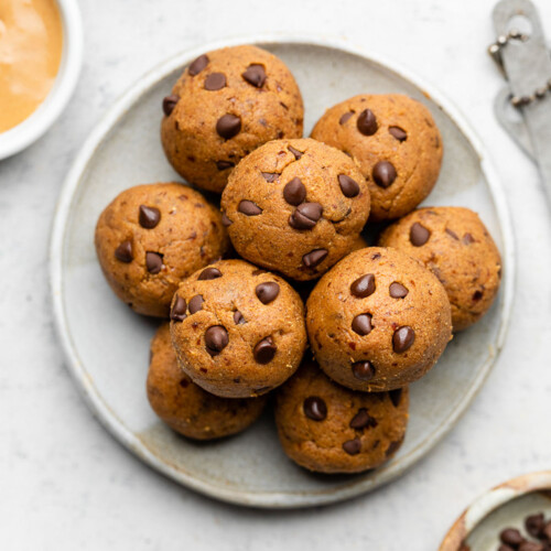 Pumpkin protein balls stacked on top of each other on a small white plate. Next to the plate is a bowl of mini chocolate chips, a bowl of nut butter, a bowl of pumpkin puree, and measuring spoons.