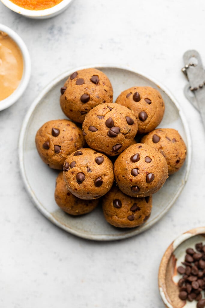 Pumpkin protein balls stacked on top of each other on a small white plate. Next to the plate is a bowl of mini chocolate chips, a bowl of nut butter, a bowl of pumpkin puree, and measuring spoons.