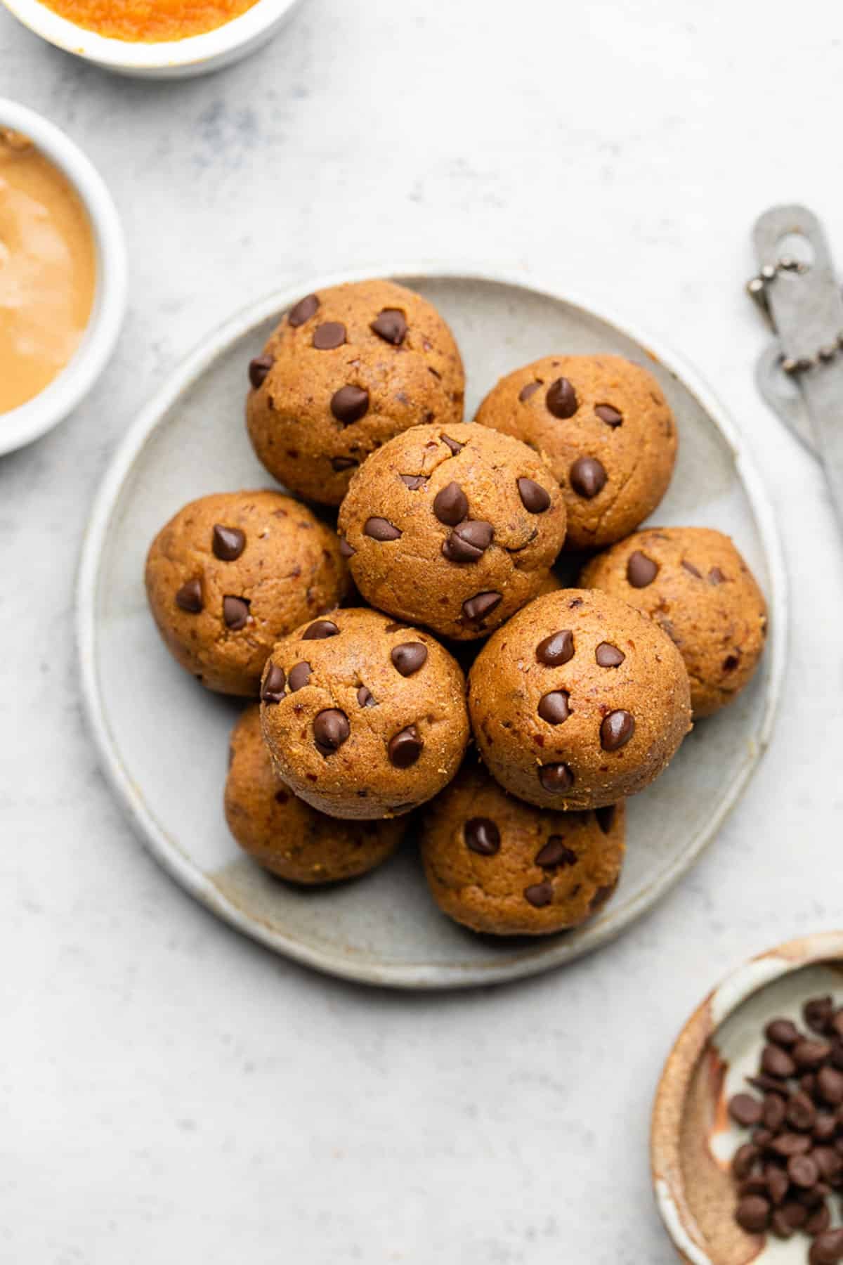 Pumpkin protein balls stacked on top of each other on a small white plate. Next to the plate is a bowl of mini chocolate chips, a bowl of nut butter, a bowl of pumpkin puree, and measuring spoons.