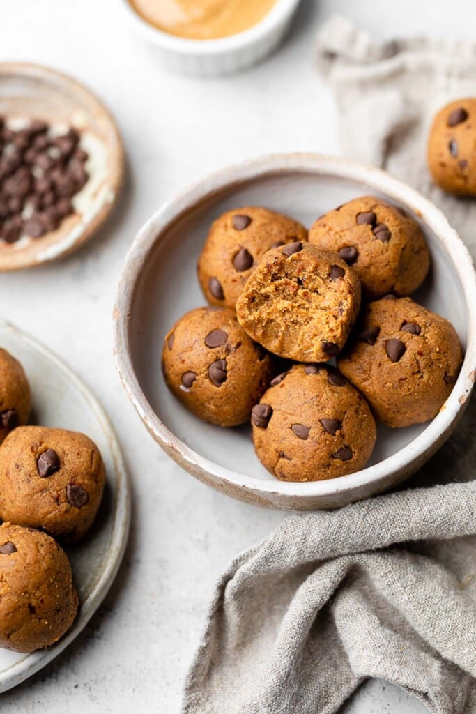 Small bowl of pumpkin energy balls stacked on top of each other. The top one has a bite taken out of it. Next to it is a plate of more energy balls, a small bowls of chocolate chips, bowl of nut butter, an energy ball by itself, and a light tan linen.