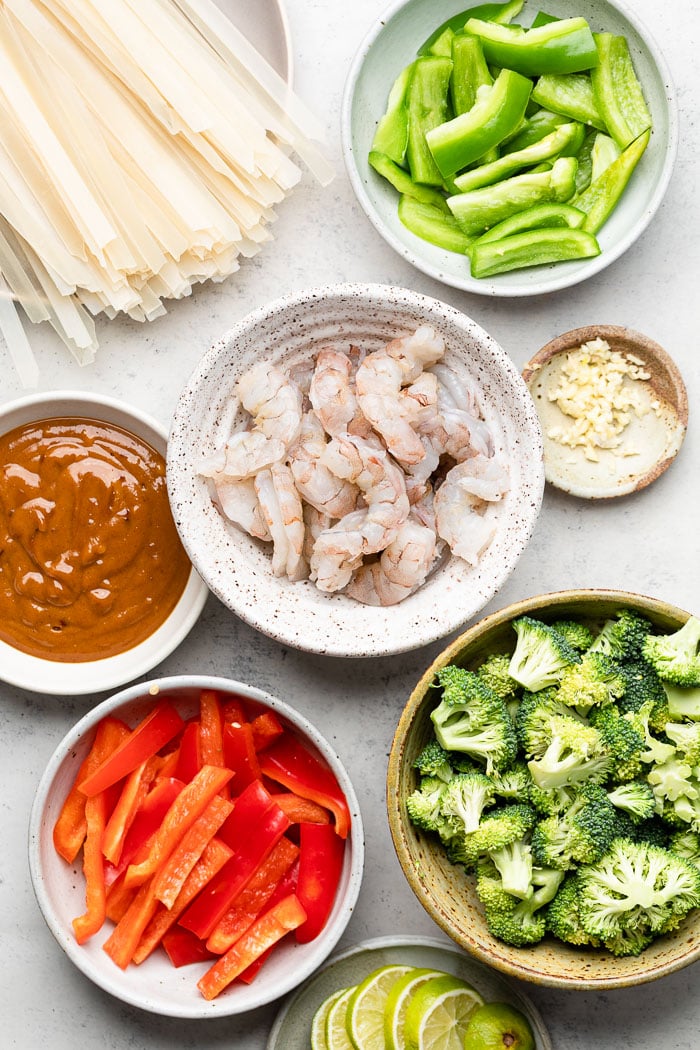 Overhead shot of bowls filled with julienned red pepper flakes, peanut sauce, raw shrimp, rice noodles, g