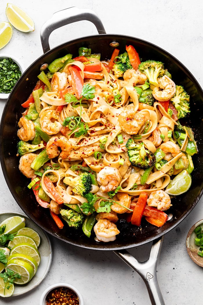 Shrimp stir fry with noodles in a large pan topped with red pepper flakes, cilantro, and green onion. Around it is a plate of lime wedges and small bowls of cilantro, green onions, and red pepper flakes.