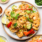 Plate of shrimp rice noodles with veggies and peanut sauce. It is topped with cilantro and has a lime wedge on the plate. Around it is another plate with shrimp noodles, small plate with lime wedges, bowl of red pepper flakes, bowl of chopped green onion, and a bowl of cilantro.