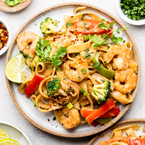 Plate of shrimp rice noodles with veggies and peanut sauce. It is topped with cilantro and has a lime wedge on the plate. Around it is another plate with shrimp noodles, small plate with lime wedges, bowl of red pepper flakes, bowl of chopped green onion, and a bowl of cilantro.