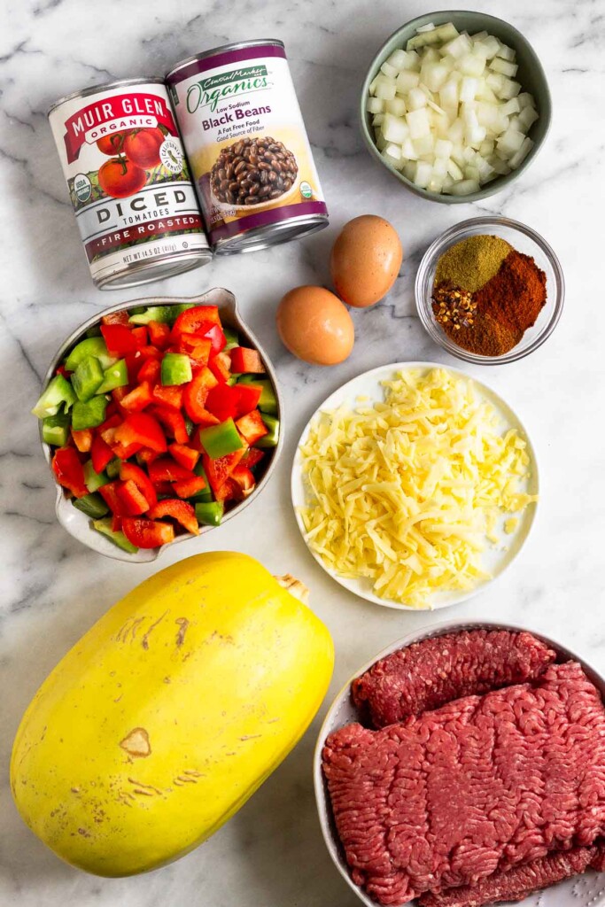 White marble counter with a bowl of diced onions, a bowl of spices, a plate of shredded cheese, a bowl of raw ground beef, a whole spaghetti squash, a bowl of diced peppers, two brown eggs, a can of roasted tomatoes, and a can of black beans.