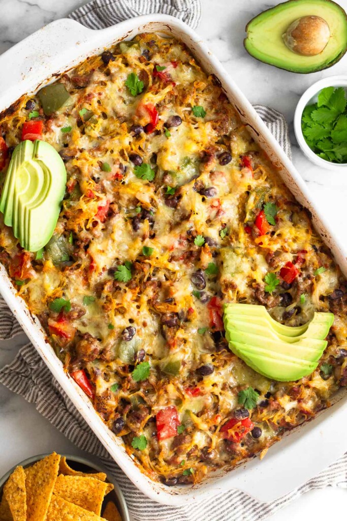 Overhead shot of chili spaghetti squash casserole in a large casserole dish. It is topped with sliced avocado and cilantro. Next to it is a bowl of corn chips, bowl of cilantro, and half an avocado.