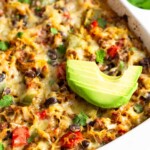 Large casserole dish with spaghetti squash casserole in it topped with sliced avocado. Behind it is a bowl of herbs and half an avocado.