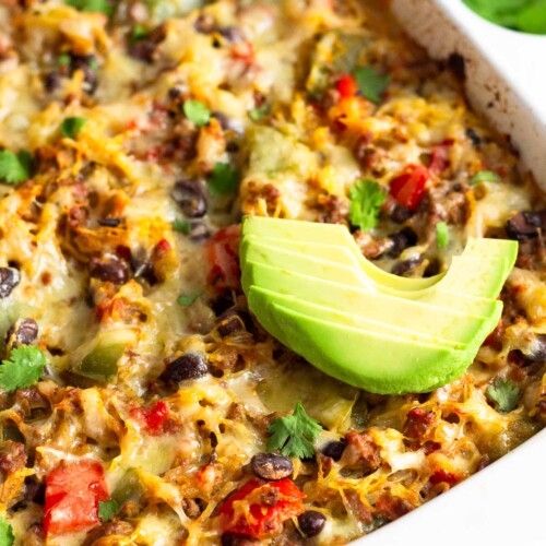 Large casserole dish with spaghetti squash casserole in it topped with sliced avocado. Behind it is a bowl of herbs and half an avocado.