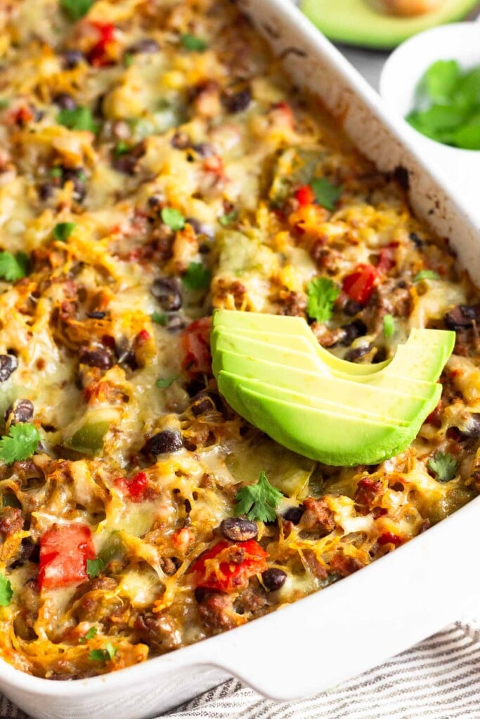 Large casserole dish with spaghetti squash casserole in it topped with sliced avocado. Behind it is a bowl of herbs and half an avocado.