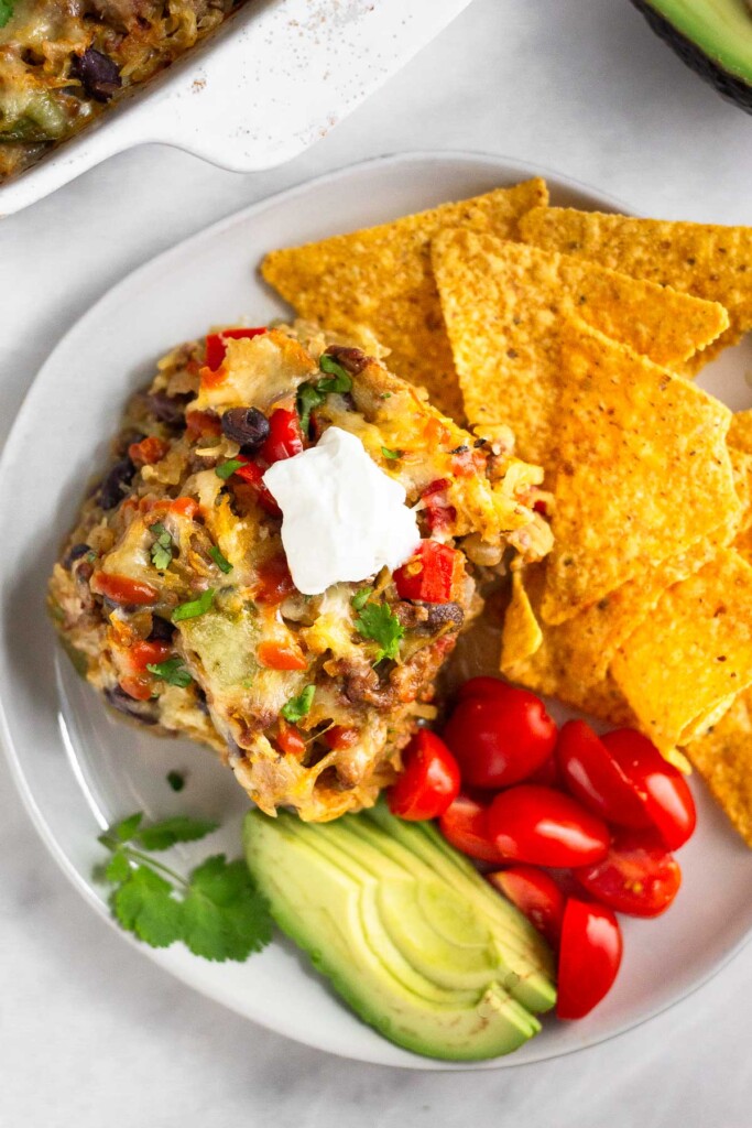A white plate with a piece of casserole, tortilla chips, diced tomatoes, sliced avocado, and cilantro. The piece of casserole is topped with greek yogurt.