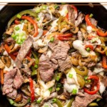 Philly cheesesteak skillet with peppers and onions and topped with melted cheese in a large pan. It is garnished with sliced green onions.