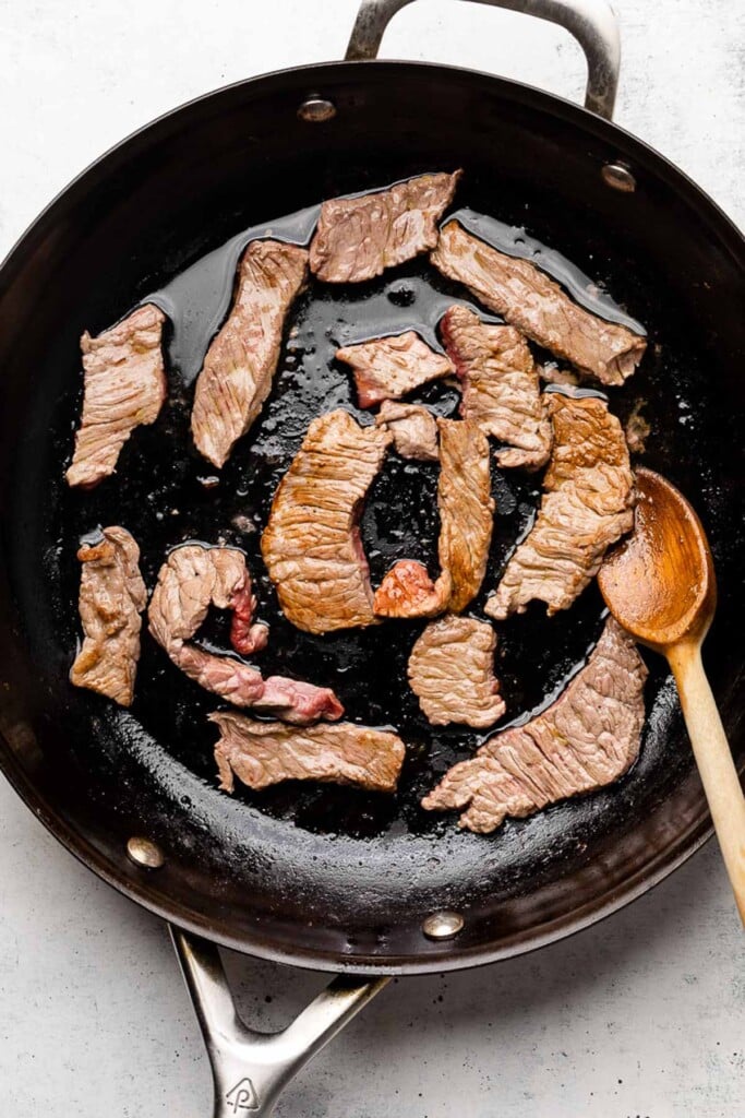 Large skillet with thinly sliced pieces of steak being cooked. A wooden spoon is also in the pan.
