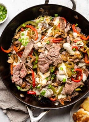 Philly cheesesteak skillet with peppers and onions and topped with melted cheese in a large pan. It is garnished with sliced green onions. Around the pan is a plate of toasted bread, a tan linen towel, and a small bowl of sliced green onions.