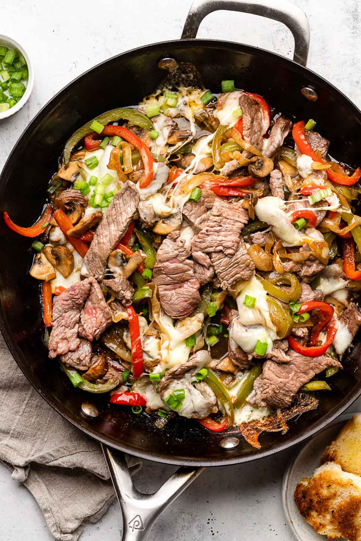 Philly cheesesteak skillet with peppers and onions and topped with melted cheese in a large pan. It is garnished with sliced green onions. Around the pan is a plate of toasted bread, a tan linen towel, and a small bowl of sliced green onions.