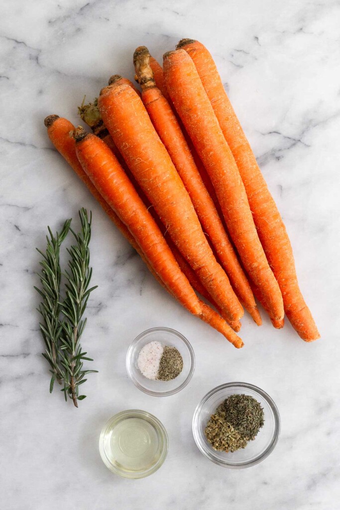 White marble counter with a pile of raw carrots, of small bowl of salt and pepper, small bowl of dried herbs, small bowl of oil, and fresh rosemary.