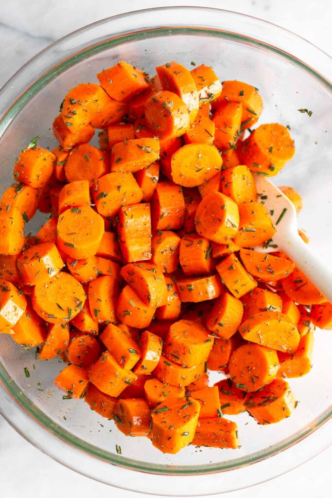 Large glass bowl filled with diced raw carrots coated in oil and herbs. There is a spatula in the bowl as well.