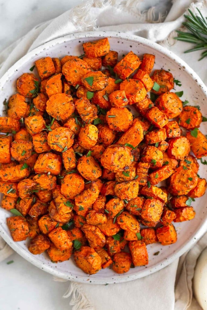 White plate filled with roasted carrots garnished with fresh herbs. Next to the plate is some fresh rosemary.
