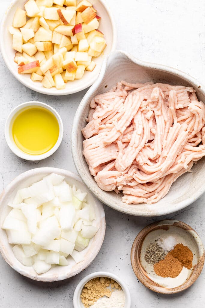 Overhead shot of a bowl of diced apple, bowl of raw ground chicken, two bowls of spices, bowl of diced onion, and a bowl of oil.