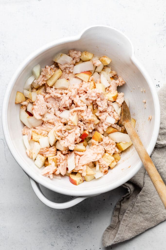 Large white bowl filled with ground chicken, diced apple, diced onion, and spices all mixed together. There is a wooden spoon in it as well.