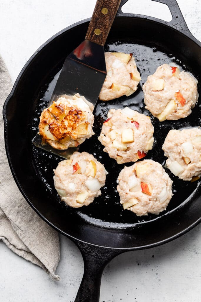 Cast iron filled with chicken apple sausage patties that are being cooked. One is being flipped over while the others are still uncooked.