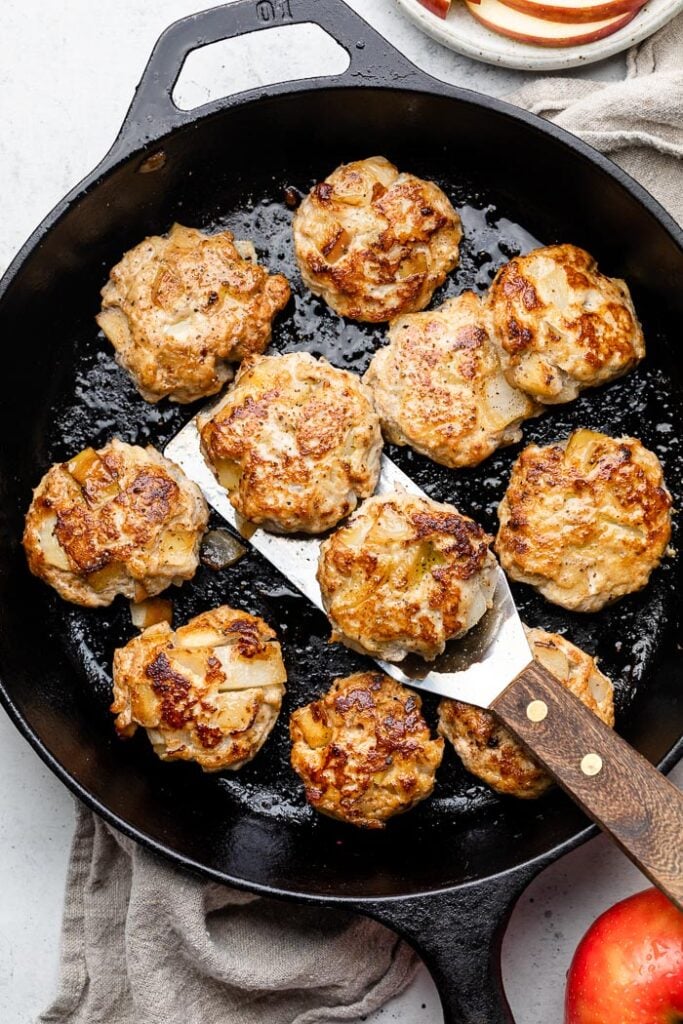Chicken apple sausage patties in a large cast iron skillet. Two of them are lifted up on a spatula. The pan is sitting a brown linen with apples around it.