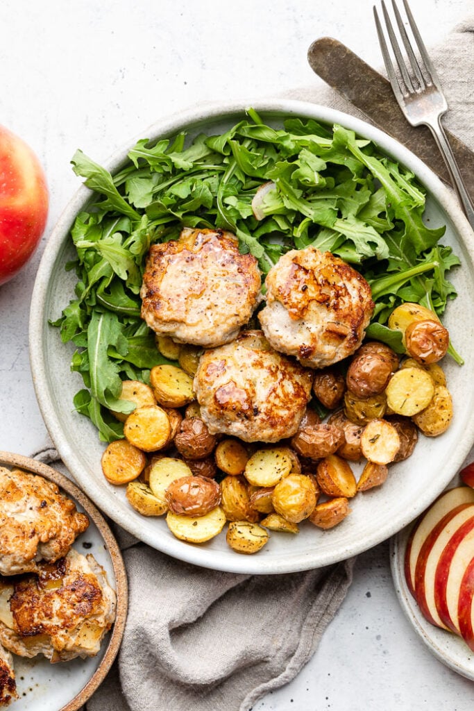 Large white plate filled with arugula, roasted baby potatoes, and three chicken sausage patties. Next to it is a small plate with more chicken sausage, an whole apple, a fork and knife, and a plate with sliced apple.