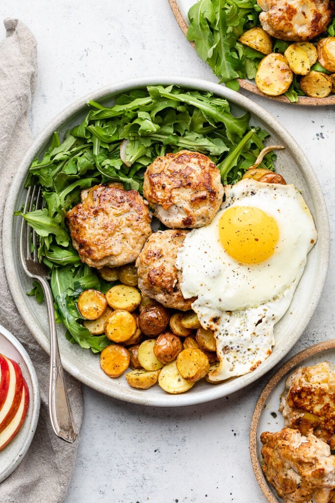 Large white plate filled with greens, roasted potatoes, chicken breakfast sausage, and a fried egg. Around the plate is a plate with sliced apples, a plate with more chicken sausage, and a plate with more greens and potatoes.