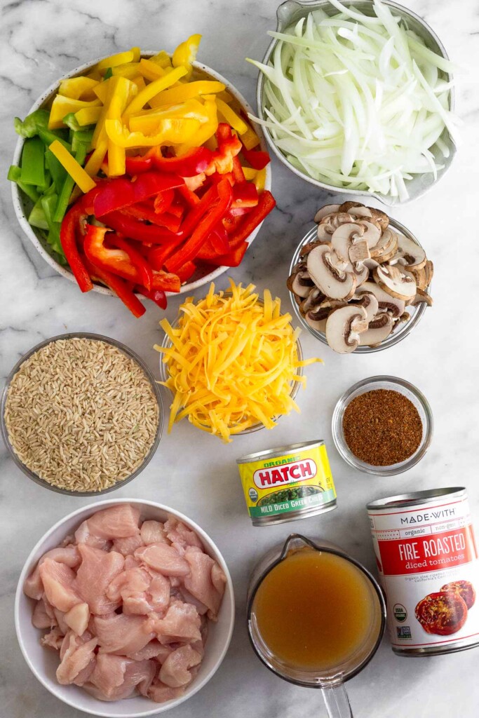 White marble counter with a bowl of julienned onions, a bowl of diced mushrooms, a bowl of taco seasoning, a can of fire roasted tomatoes, a measuring cup of broth, a can of hatch chili peppers, a bowl of raw diced chicken, a bowl of uncooked brown rice, a bowl of shredded cheese, and a bowl of julienned bell peppers.