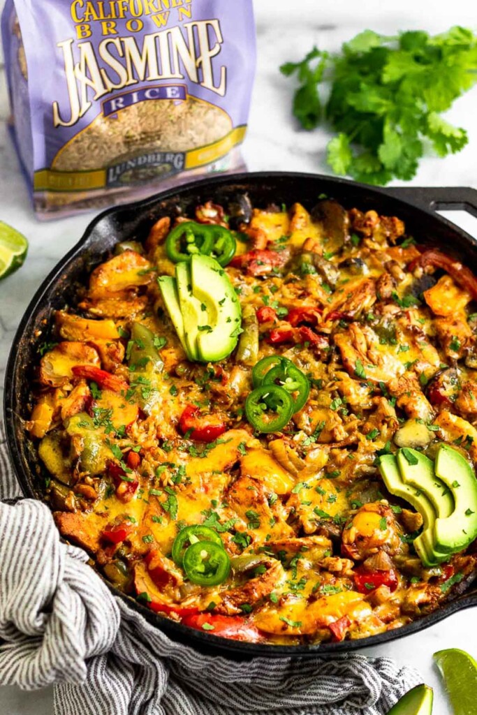 Cast iron skillet filled with fajita chicken casserole topped with melted cheese, avocado, sliced jalapeños, and cilantro. Behind the pan is a bag of brown rice and a bunch of cilantro.