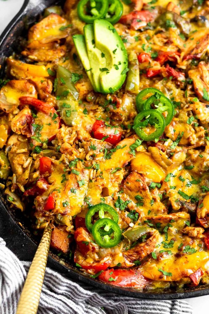 Fajita chicken casserole topped with melted cheese and garnished with avocado, jalapeño slices, and cilantro in a large pan. A gold spoon is in the pan.