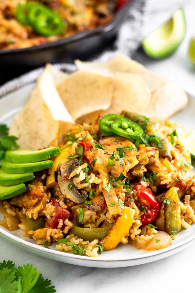 White plate filled with chicken fajita casserole, sliced avocado, and some tortilla chips. Behind it is a cast iron skillet with more casserole and a piece of avocado.