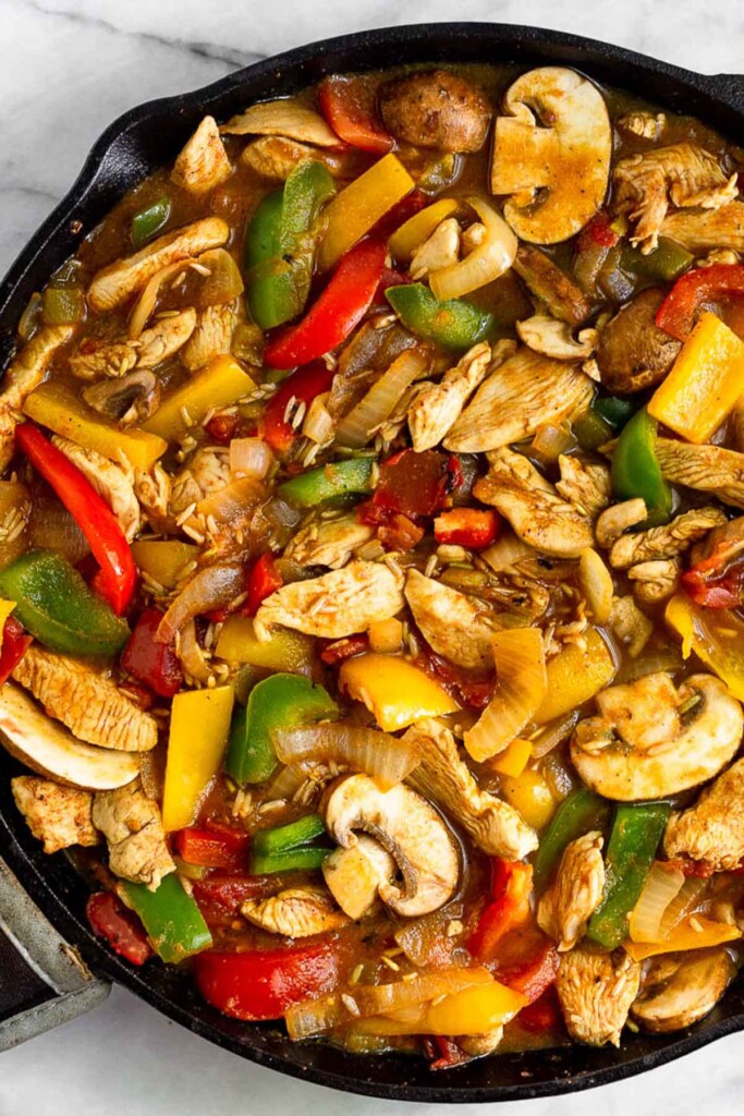 Overhead shot of a large cast iron skillet filled with sautéed chicken, sautéed bell peppers, sautéed onions, mushrooms, uncooked rice, and broth.